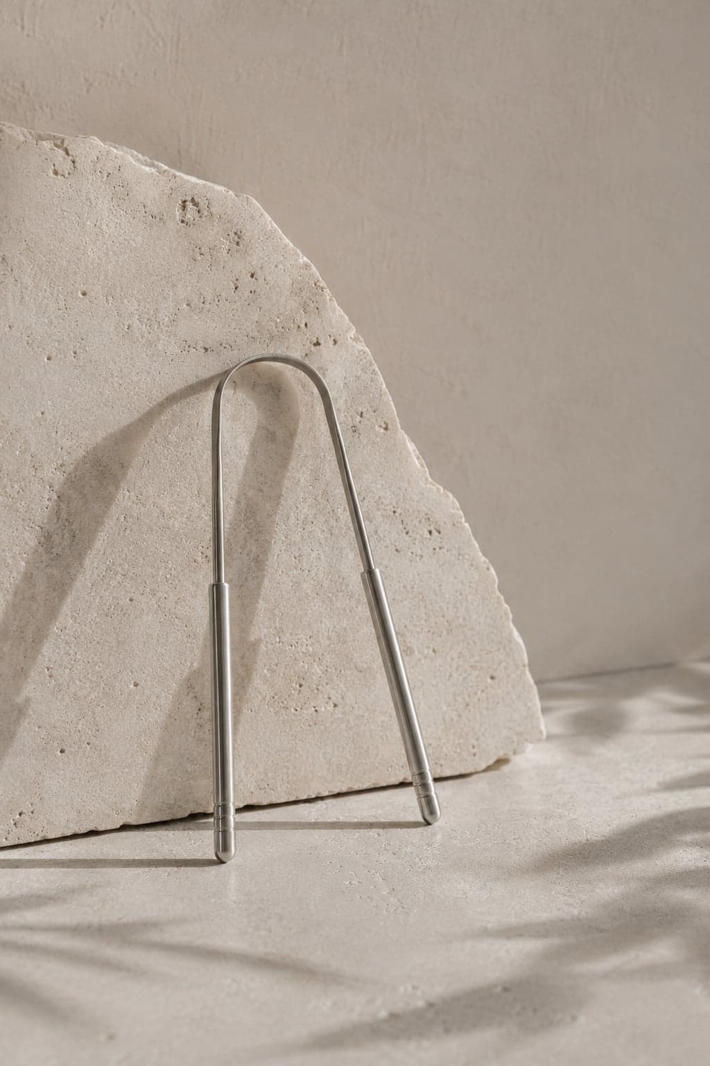 Tongue Scraper No. 01 — brushed stainless steel, leaning against travertine